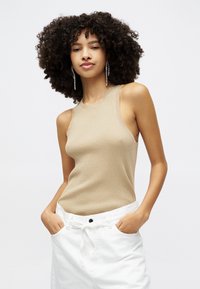 Beige ribbed tank top dengan leher bulat dan desain tanpa lengan, dipadukan dengan celana putih yang tinggi di pinggang. Latar belakang berwarna putih polos.