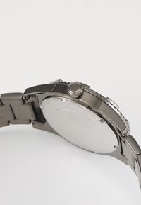 Rustfritt stål klokke med rund kasse og strukturert bezel. Har en fremtredende krone og en polert bakside som viser gravering detaljer.