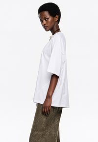 Camiseta blanca de gran tamaño con mangas cortas, cuello redondo y aberturas laterales, combinada con pantalones verde oliva texturizados que tienen bolsillos.