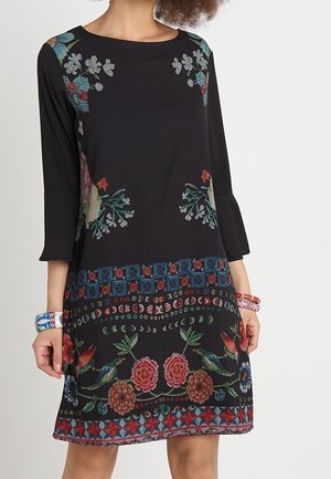 Frau trägt ein schwarzes Kleid mit dreiviertel Ärmeln, das mehrfarbige Blumen- und Vogelmuster sowie geometrische Bordüren am Saum und an der Taille aufweist.