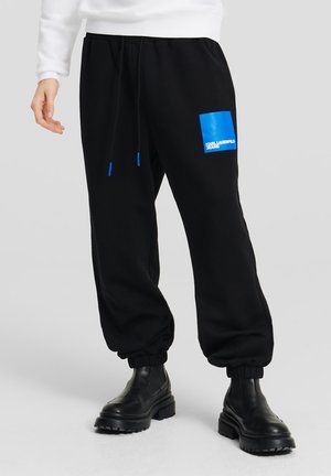 Sorte sweatpants med et blåt rektangulært grafisk design på venstre ben, elastiske manchetter og snor med blå spidser.