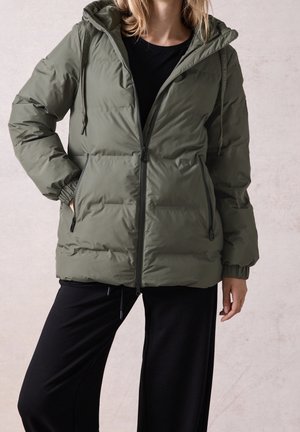 Woman wearing a green hooded puffer jacket zipped up, with hands in pockets, paired with black pants against a plain light background.