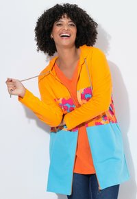 Felpa con zip colorata in arancione, blu e tessuto fantasia; presenta un cappuccio con coulisse e maniche lunghe; indossata sopra una maglia arancione.