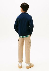 Giovane con le mani in tasca, indossando una giacca blu navy, una camicia a quadri verde, pantaloni cargo beige e sneaker beige su sfondo bianco.