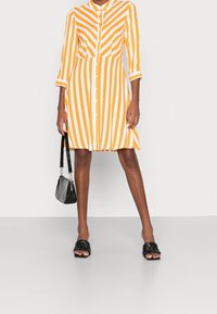 Vestido a rayas en colores naranja y blanco, con botones en la parte delantera, cuello, mangas de tres cuartos y un corte holgado. Usado con un bolso negro y sandalias.