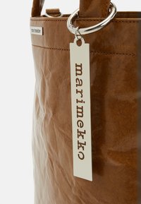 Sac marron texturé avec des coutures surpiquées et une étiquette métallique argentée suspendue à un grand anneau en argent, affichant le nom de la marque Marimekko.
