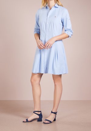 Robe chemise bleu clair avec un devant à boutons, des manches longues et une jupe plissée. Associée à des sandales à talons blocs bleu foncé.