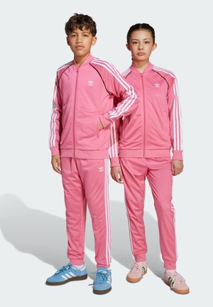Zwei Personen tragen passende rosa Trainingsanzüge mit weißen Streifen und schwarzen Logoakzenten. Die Outfits haben ein Reißverschlussdesign und Taschen.
