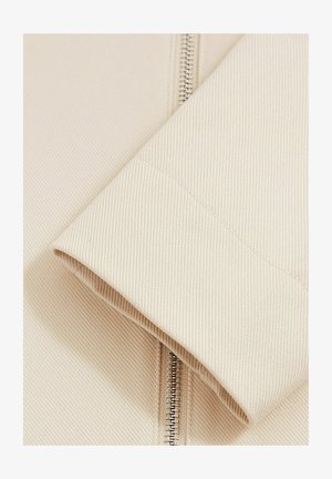 Beige tekstuuriinen kangasslevy, jossa on näkyvä vetoketju, esittelee uritetun kuvion ja siistiä ommelta. Tasainen pinta, jossa on pieni kiilto.