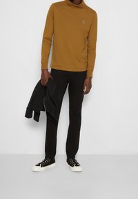 Senfgelbes Rollkragen-Shirt mit langen Ärmeln; kombiniert mit schwarzen Hosen und einer schwarzen Jacke; getragen mit schwarz-weißen Sneakers.