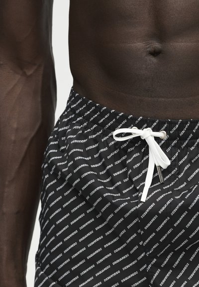 Torse musclé d'une personne à la peau foncée portant un short de bain noir avec un motif de texte blanc "DSQUARED2" et un cordon blanc à la taille.