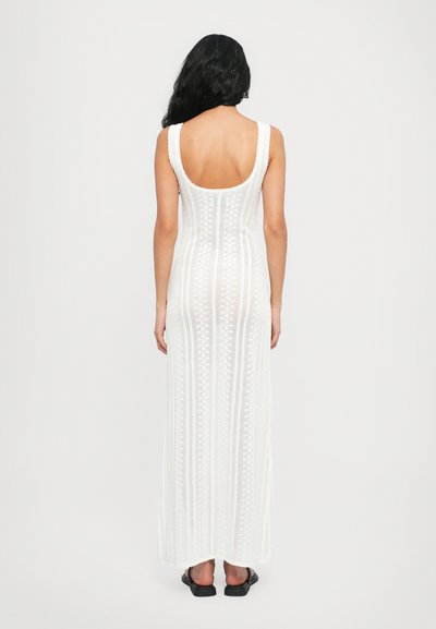 White, knitted maxi dress with a scoop back, featuring a textured, patterned design. The model wears black sandals.
