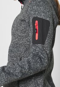 Veste en polaire gris foncé avec une finition texturée, dotée d'un patch noir sur la manche et d'une fermeture éclair rouge sur la poche, avec une coupe ajustée.