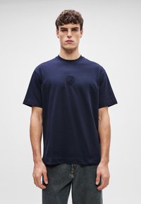 Ναυτικό μπλε βαμβακερό μπλουζάκι με στρογγυλή λαιμόκοψη, κοντά μανίκια και υφή με λογότυπο "B" στο στήθος, συνδυασμένο με σκούρα τζιν.