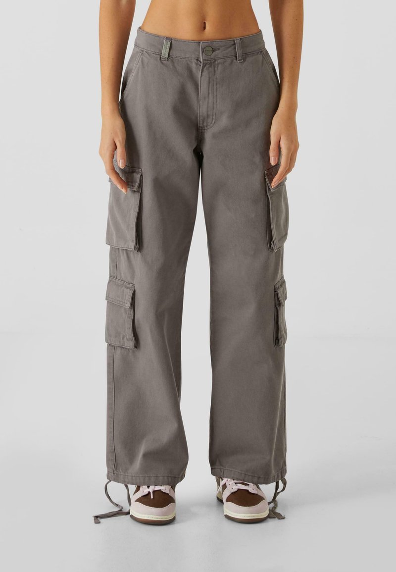 Pantalons cargo en tissu gris avec une coupe ample, dotés de plusieurs poches latérales et de ourlets à cordon de serrage. Fermeture à bouton et à fermeture éclair à la taille.
