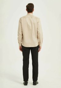 Chemise beige à manches longues avec un col boutonné, associée à un pantalon et des chaussures noirs. Texture lisse, design simple, vue arrière montrée.