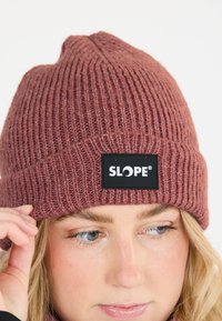 Ribbad stickad mössa i dammig ros, med en svart lapp med "SLOPE"-logotyp. Mjukt material, åtsittande design med en spets på toppen.