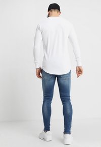 Vit långärmad skjorta med figurnära stil, i kombination med smala blå denimjeans. Skorna är vita sneakers. Utsikt från baksidan.