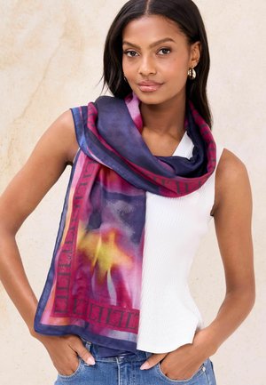 Žena s farebným hodvábnym šálom v odtieňoch fialovej, ružovej a žltej, obtočeným okolo bieleho tielka bez rukávov a modrých džínsov.