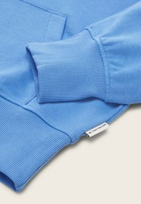 Sudadera azul claro con puños y dobladillo acanalados, textura suave y una etiqueta de marca visible con "TOM TAILOR" en la parte inferior derecha.