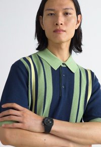 Polo à manches courtes en marine et vert avec des rayures verticales, comportant un col vert clair et une patte de boutonnage à trois boutons. Montre noire au poignet.