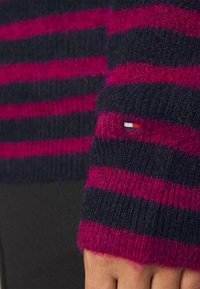 Sweter robiony na drutach, w czarno-fuksjowe paski, wykonany z miękkiego, puszystego materiału. Zawiera mały, haftowany logo na mankiecie.