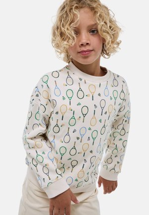Enfant aux cheveux bouclés blonds portant un sweat-shirt crème orné d’un motif de raquettes de tennis colorées et de volant de badminton, debout devant un fond blanc.