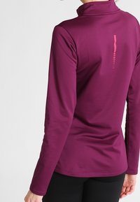 Långärmad topp i mörk plommon, tillverkad av ett mjukt tyg. Har en hög krage och en minimalistisk grafisk design i rosa på baksidan.