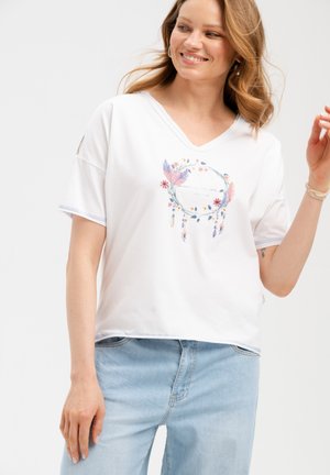 Uśmiechnięta kobieta w białym T-shircie z dekoltem w serek, z kolorowym kwiatowym wzorem łapacza snów, oraz jasnoniebieskich dżinsach na jednolitym tle.