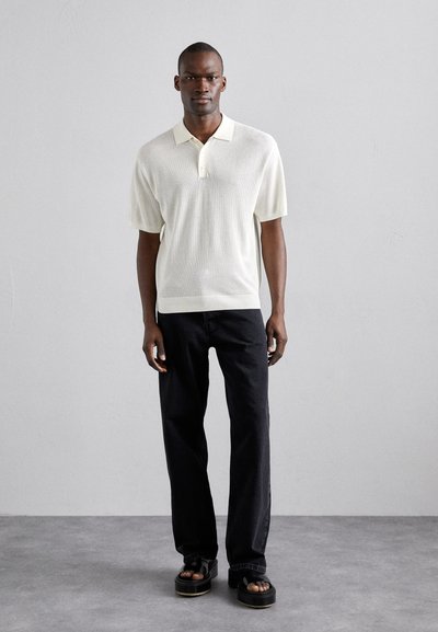 Polo à manches courtes en tissu léger crème, avec un col et une patte de boutonnage, assorti à un jean noir à jambes droites et des sandales à plateforme.