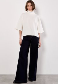Weißes Turtleneck-Oberteil mit Waffelstruktur und weiten Ärmeln, kombiniert mit hoch taillierten, dunklen Jeans mit weitem Bein und sichtbaren Nähten.