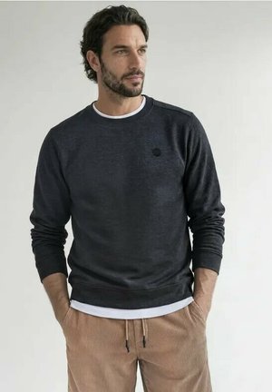 Man met donker krullend haar en baard, die een donker shirt met lange mouwen en lichtbruine corduroy broek draagt, staat met handen in de zakken.