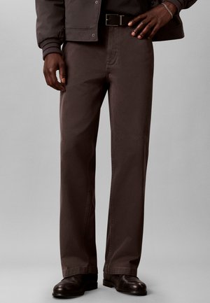 Mann trägt braune Hose mit geradem Bein und schwarzem Gürtel, dunkelbraune Lederschuhe und dunkle Jacke, steht vor grauem Hintergrund.