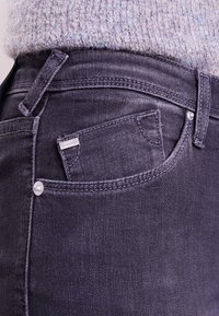 Ciemnoszare dżinsy mają małą przednią kieszeń, przeszycia oraz metalowy guzik na pasku.