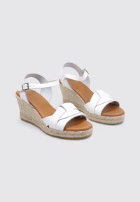 Sandalias de cuña blancas con tiras cruzadas, plantilla en tono bronceado y base tejida estilo alpargata. Correa ajustable en el tobillo con hebilla metálica.