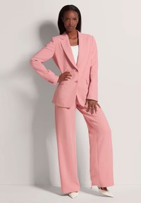 Roze op maat gemaakt pak bestaande uit een lange blazer met gelapte revers en brede broekspijpen, gecombineerd met een witte top en puntige hakken.