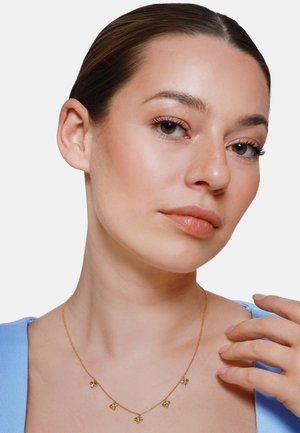 Donna con capelli raccolti che indossa un top azzurro chiaro e una collana d'oro con cinque piccoli ciondoli a forma di ape.