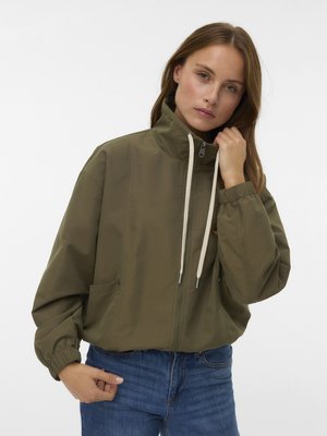 Femme ajustant le col d'une veste zippée vert olive avec cordons, portant un jean bleu sur un fond blanc uni.