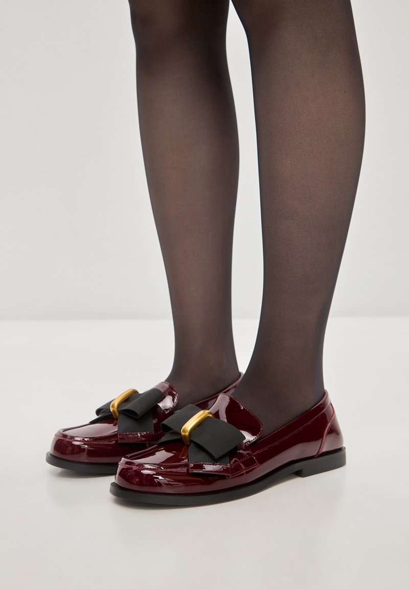 Mocassins en cuir verni bordeaux avec un grand nœud noir et des détails en métal doré, présentant une silhouette élégante et une semelle plate noire.