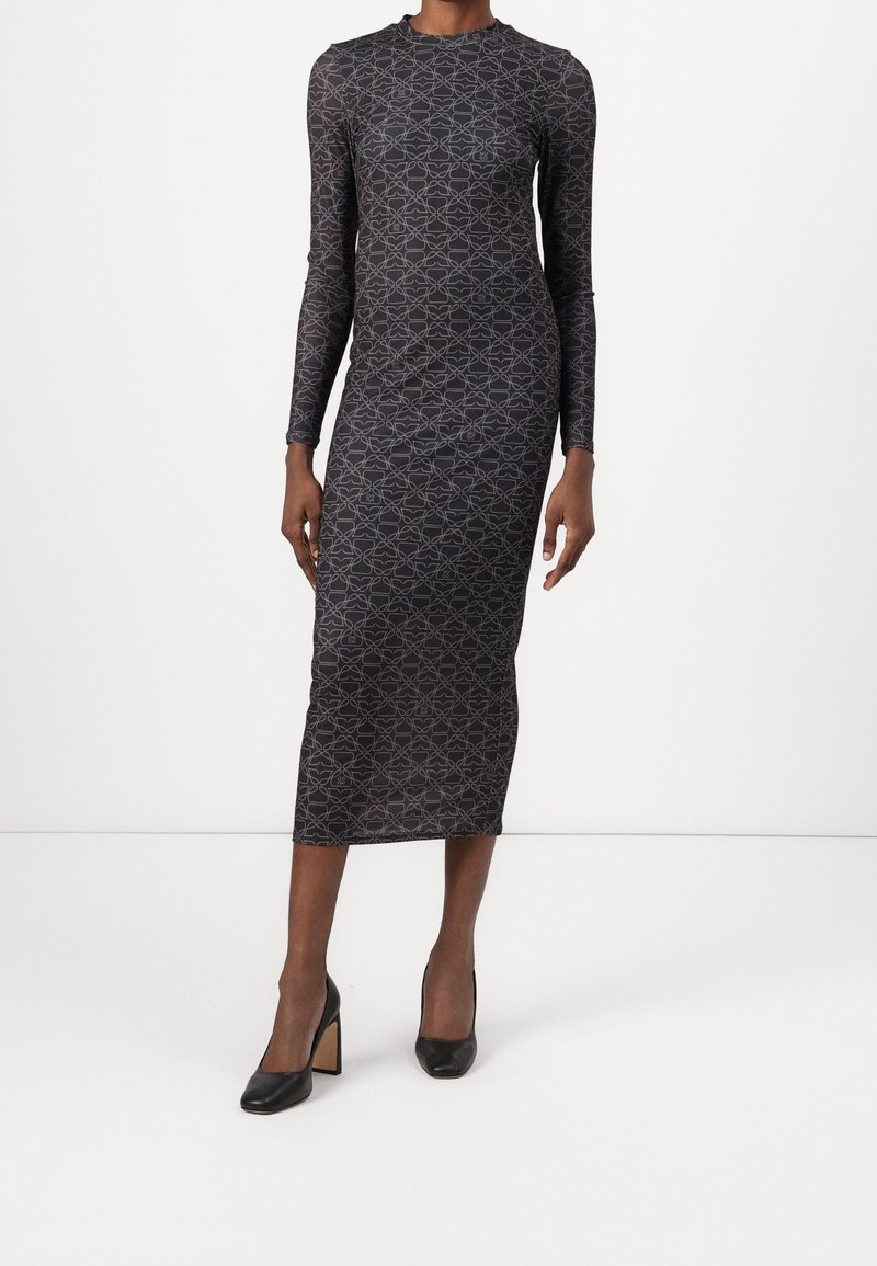 Femme portant une robe midi noire ajustée à manches longues avec un motif géométrique gris clair complexe, associée à des talons hauts noirs.