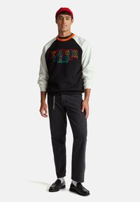 Czarna bluza z białymi rękawami, logo "STRANGER THINGS" w kolorowym napisie, zestawiona z ciemnymi dżinsami i czarnymi butami. Czerwona czapka na głowie.