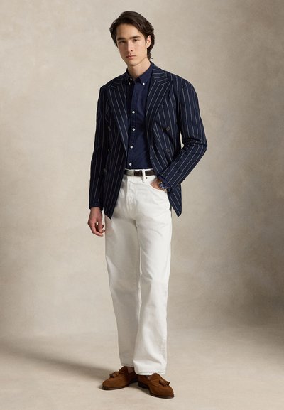 Jaunas vyras stovintis dėvi tamsiai mėlyną pinstripe švarką, tamsiai mėlyną marškinius, baltas kelnes, rudas tassel loafers, viena ranka kišenėje.