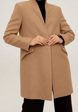 Vrouw die een camelkleurige jas met één knoop sluit over een zwarte coltrui en broek, staand tegen een effen lichte achtergrond.