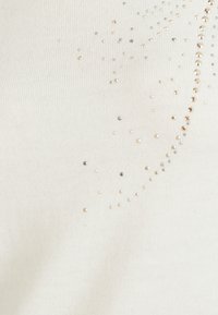 Witte top met korte mouwen, gemaakt van een textuurstof voorzien van verspreide metalen versieringen in goud en zilver, wat een stippenpatroon creëert.