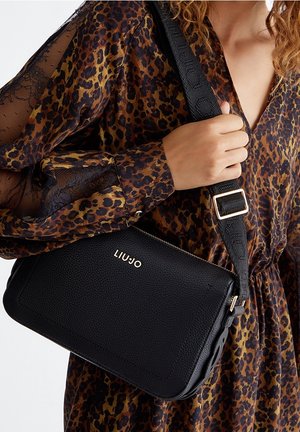 Kobieta w sukience w panterkę trzyma czarną torebkę Liu Jo na ramię z szerokim paskiem i złotą klamrą.