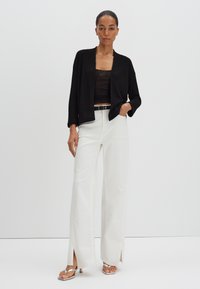 Zwarte geribbelde cardigan over een zwart hemdtop, gecombineerd met brede witte broeken met zijsplitten; model draagt zilveren sandaal met bandjes.