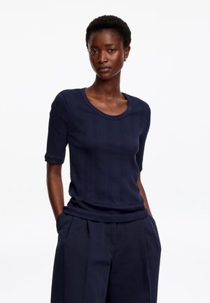 Femme aux cheveux courts et bouclés portant un haut à manches courtes côtelé bleu marine et un pantalon bleu marine, debout les mains dans les poches sur fond blanc.