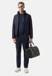 Marineblå quilted jakke, mørkeblå sweatshirt, matchende bukser og hvide sneakers. Modellen holder en sort læder duffeltaske. Blå solbriller.