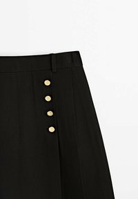 Jupe noire taille haute, quatre boutons décoratifs dorés alignés verticalement sur le côté gauche, et une passants de ceinture à la taille.