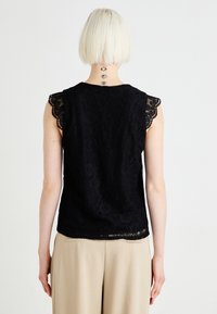 Svart blonde overdel med korte ermer og intrikate blomster mønstre, kombinert med lyse beige bukser. Bakvisningen viser bølgete kanter.